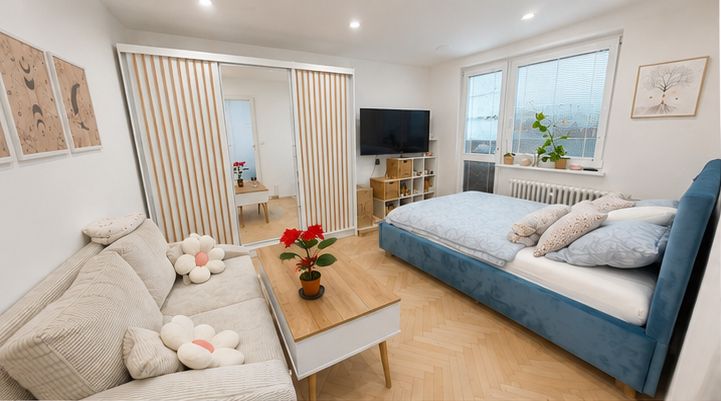 Heller Wohn- und Schlafraum mit Fischgrätparkett, blau gepolstertem Doppelbett, Sofa, Couchtisch und Schrank mit Holzlamellen.
